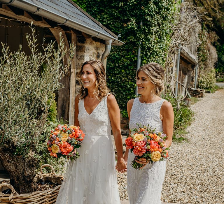 Brides walk together outdoors whilst holding bright floral bouquets and wearing boho chic wedding gowns