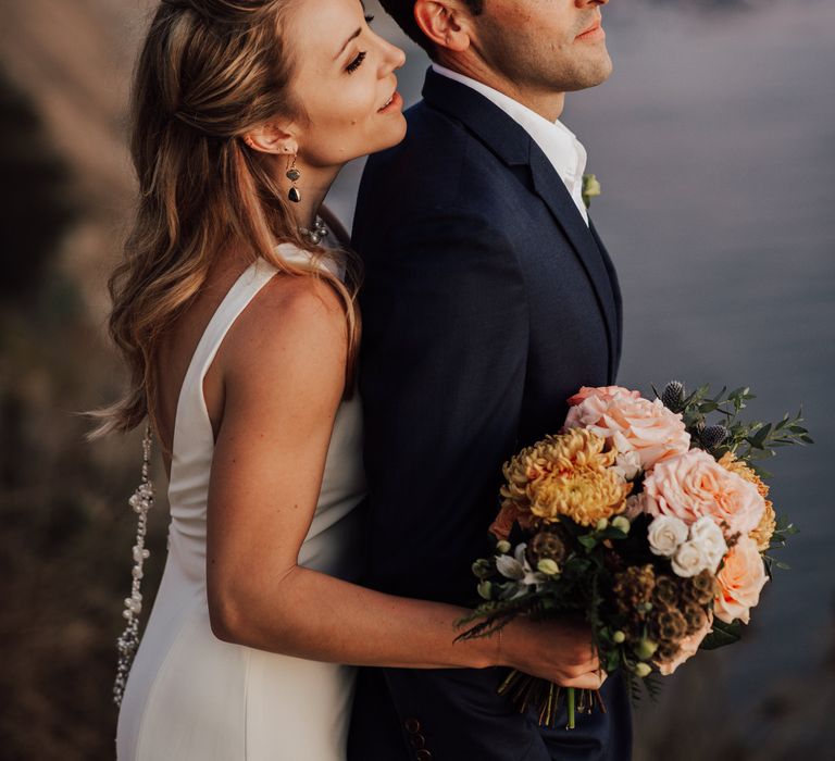 Blonde bride embraces groom as she wraps her arms around him and they look out across the ocean