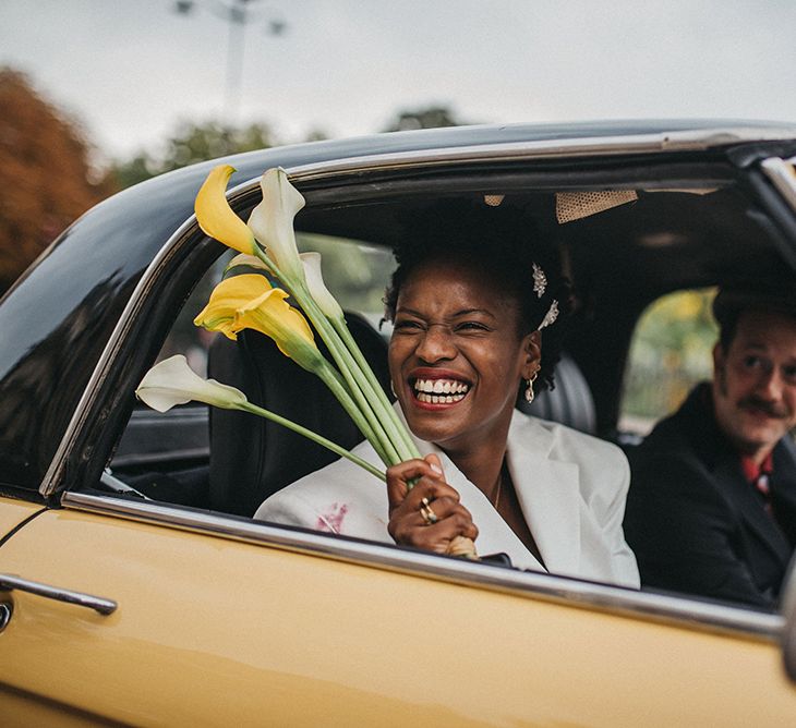 A Black bride laughs out of the window of a yellow car. She holds lilies in her hand,