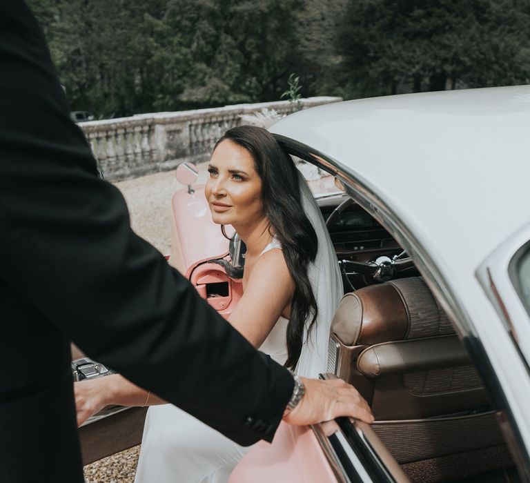 Bride with long dark curled wedding hair wearing white Pronovias wedding dress and veil gets out of pink Cadillac whilst looking up at groom in black tuxedo outside of Came House Dorset