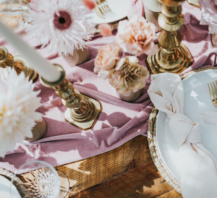 Place setting with natural wicker place mat crackled tableware and gold cutlery surrounded by gold candlesticks and blush flowers in inkwells 