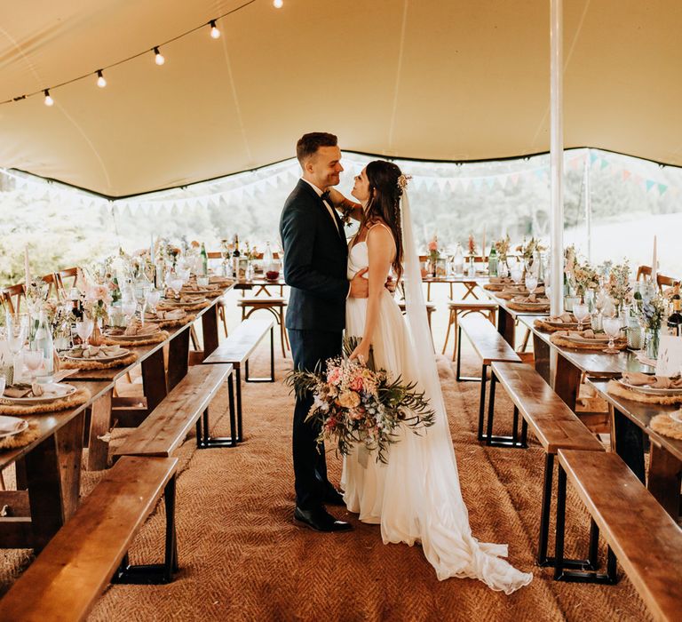 Bride in white cami wedding dress and train holding multicoloured bridal bouquet smiles at groom in blue suit and bow tie in tipi with strung fairy lights and wooden tables at garden wedding reception