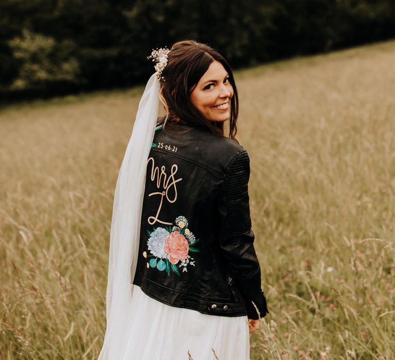 Bride in white dress and floor length veil stands smiling in field whilst wearing hand painted leather bridal jacket at garden wedding reception