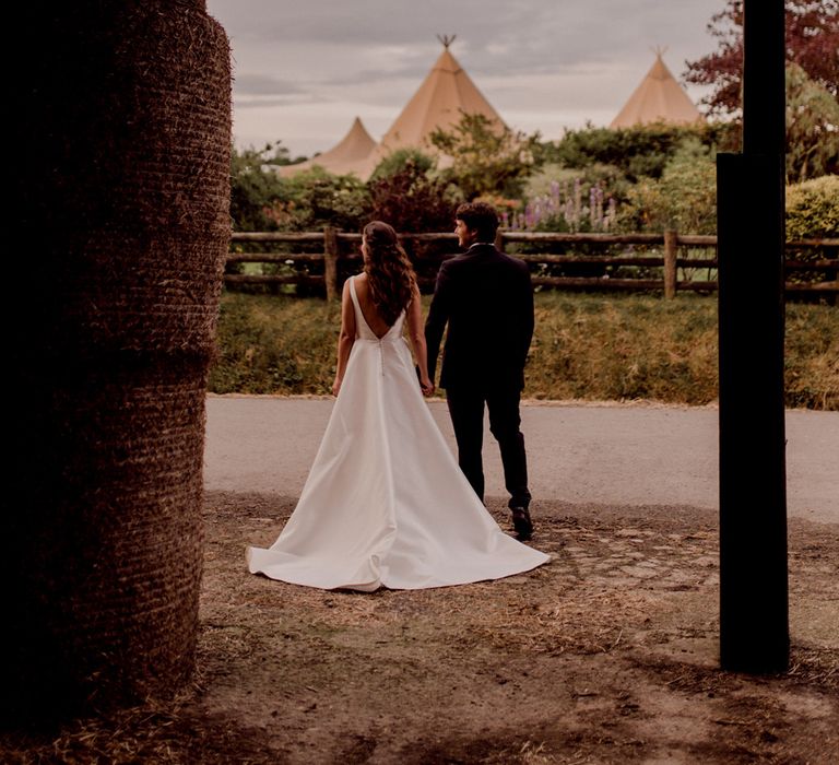 Bride in white low back Elbeth Gillis gown and long curled hair stands holding hands with groom in dark brown suit outside barn at home farm wedding with tipi