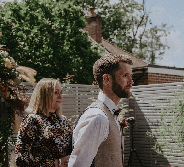 Groom with beard wearing light brown waistcoat, bow ties and dried flower buttonhole and celebrant in long sleeved patterned wedding dress watch bride come up the aisle for garden wedding with burnt orange wedding theme  