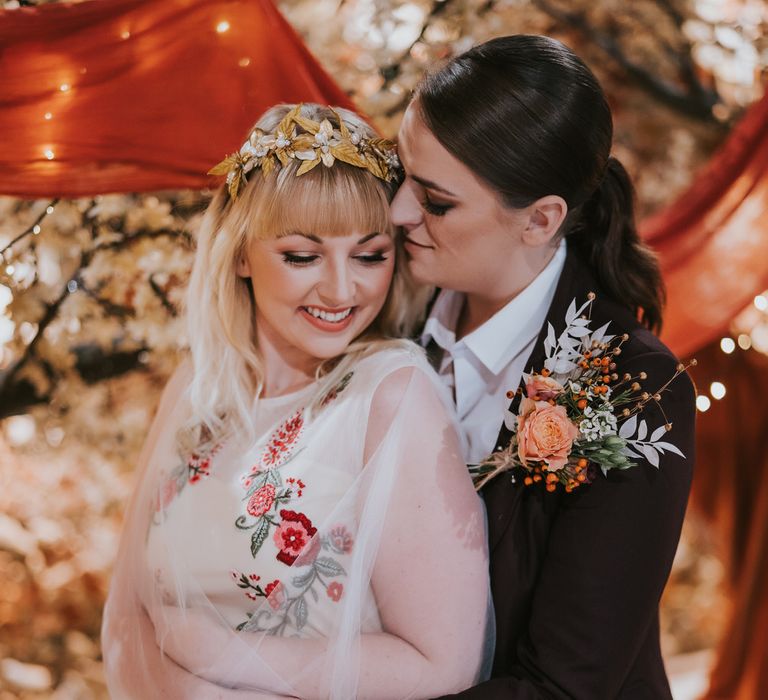 Two brides hugging at pumpkin spice wedding altar, one bride is wearing a wedding dress with red flower embroidery detail, and mesh cape veil, the other is wearing a plum coloured suit