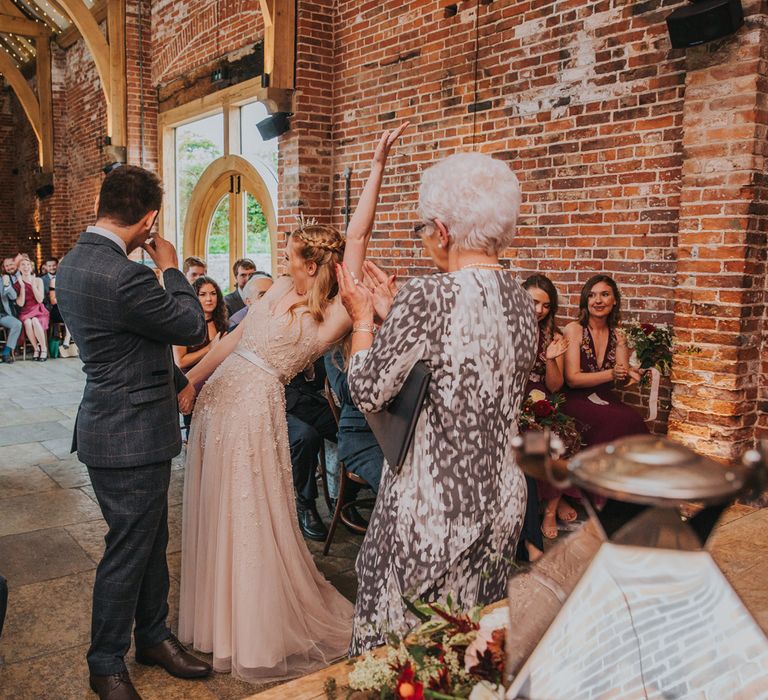 Bride in pearl wedding dress throws hands in the air with joy standing with groom in checked suit during barn wedding ceremony