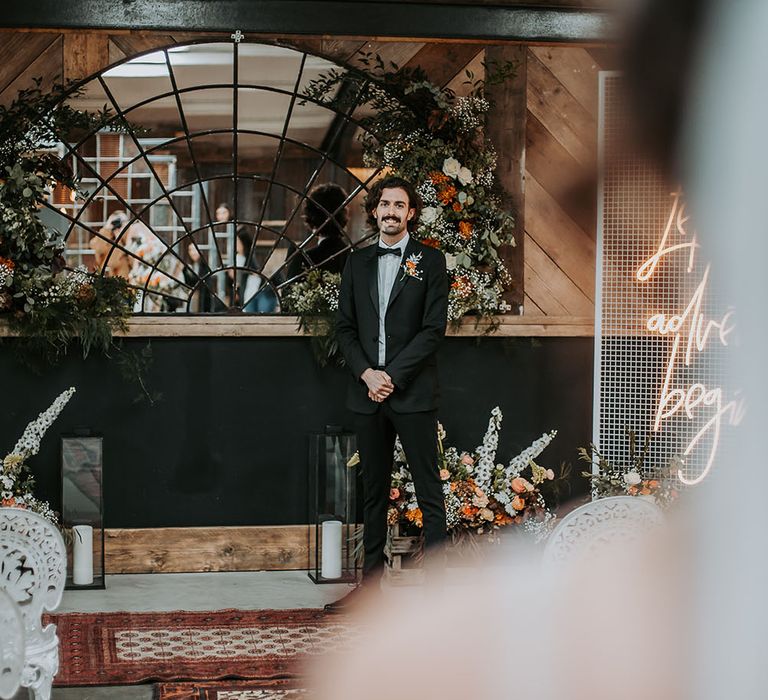 Groom in a black tuxedo with orange buttonhole flower standing at the altar of rustic Lake District wedding venue 