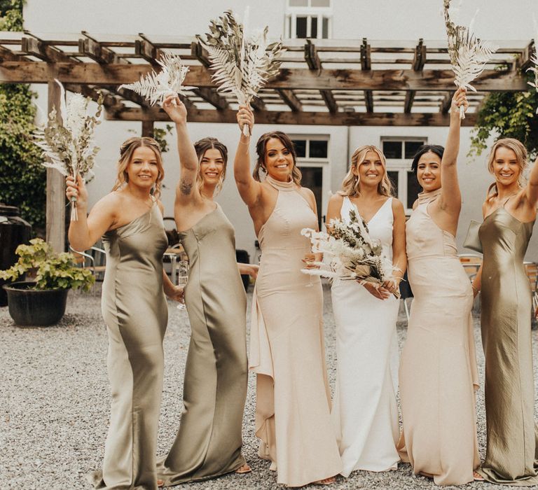 Bride in white V neck Made With Love wedding dress holding white and green dried bridal bouquet stands with bridesmaids in satin sage green and high necked beige bridesmaids dresses whilst they wave fern bridesmaids bouquet at Anran Devon