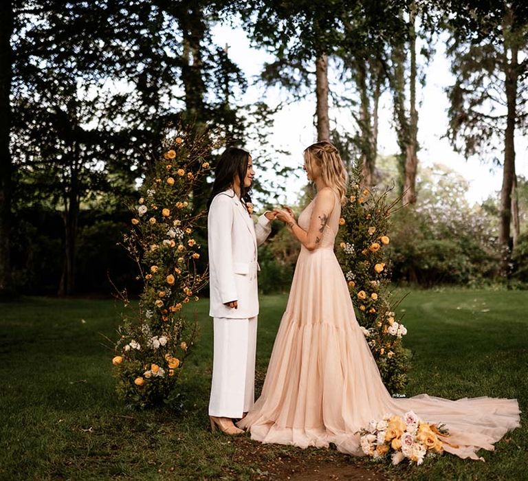 Outdoor wedding ceremony with two brides in a white suit and blush pink dress exchanging vows 