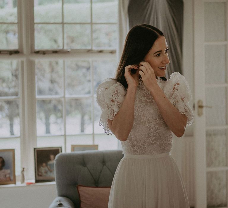 Bride in white lace top puffed sleee Daalarna wedding dress puts in her earrings before Surrey summer wedding