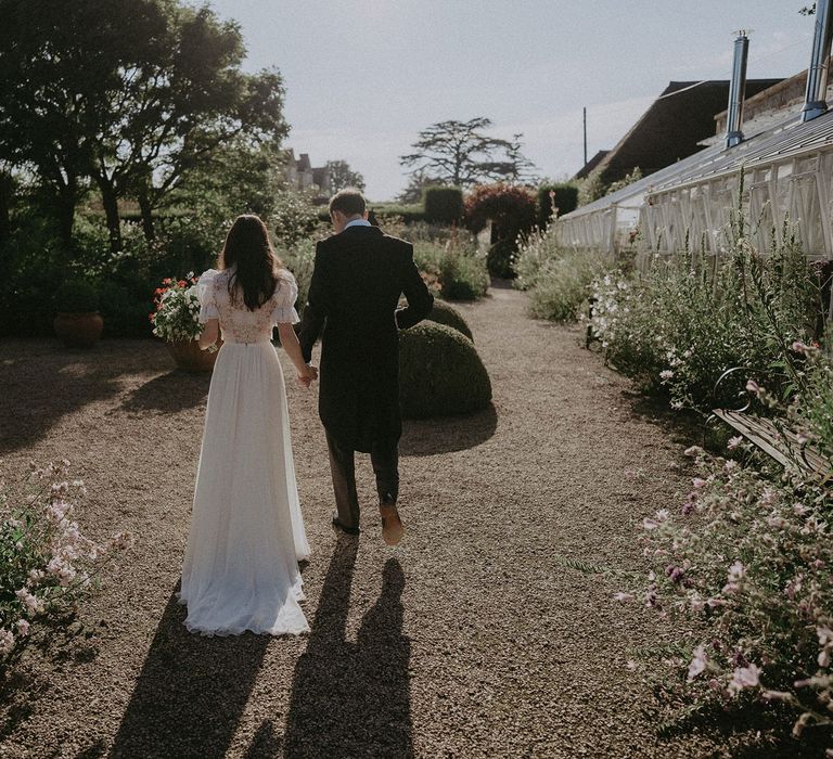 Bride in white lace puffed sleeve Daalarna wedding dress walks through grounds of Loseley Park holding hands with groom in morning coat after wedding