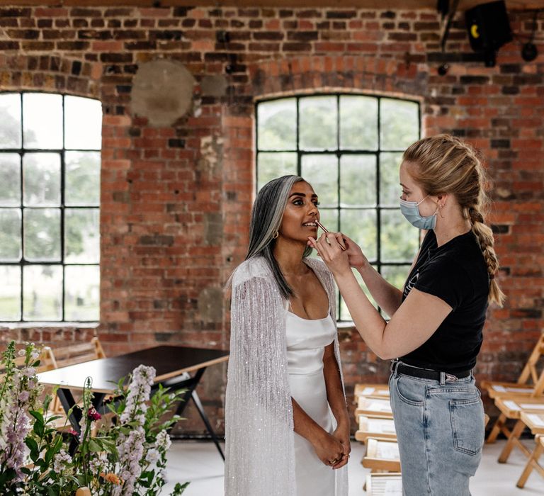 Bride in satin Halfpenny London wedding dress and tasselled bridal cape gets her make up done by woman in jeans and black top before wedding at Loft Studios London