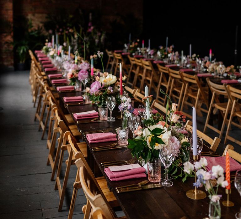 Long wooden table with folding wooden chairs, multicoloured candles, mixed florals and pink napkins in industrial room with festoon lighting for wedding breakfast at Loft Studios London