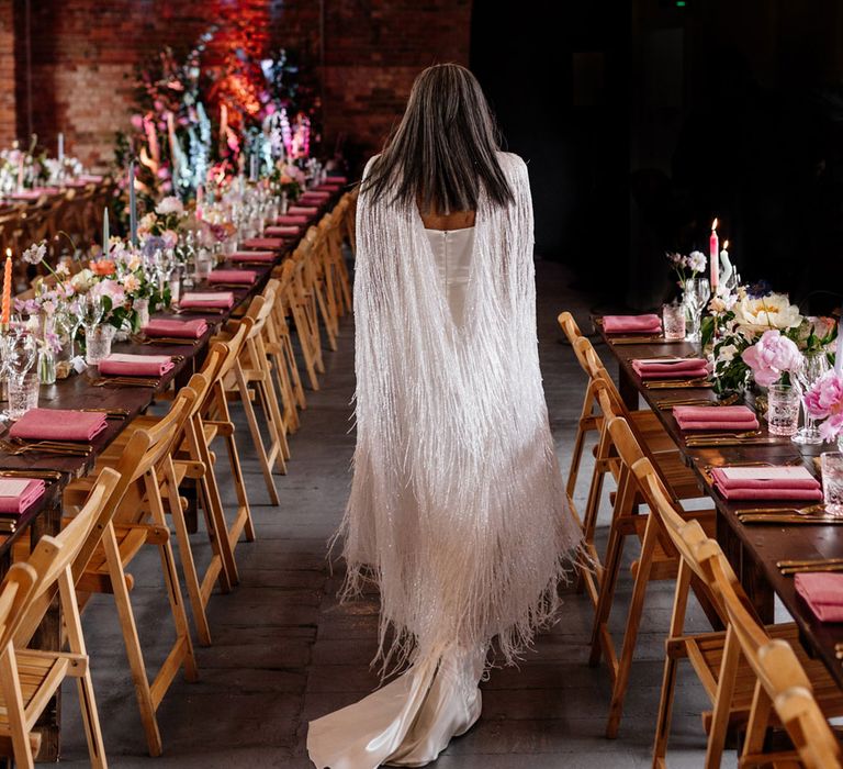 Bride in Halfpenny London tasselled bridal cape walks in-between long wooden wedding breakfast tables laid with candles, florals and pink napkins in industrial room with wooden brans and festoon lighting at Loft Studios London