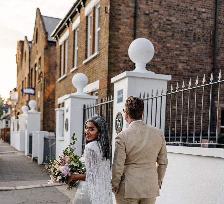 Bride in satin Halfpenny London wedding dress and tasselled bridal cape holding multicoloured bridal bouquet looks over her shoulder as she holds hands with groom in linen suit and gold Nike trainers as they walk down the street during wedding at Loft Studios London