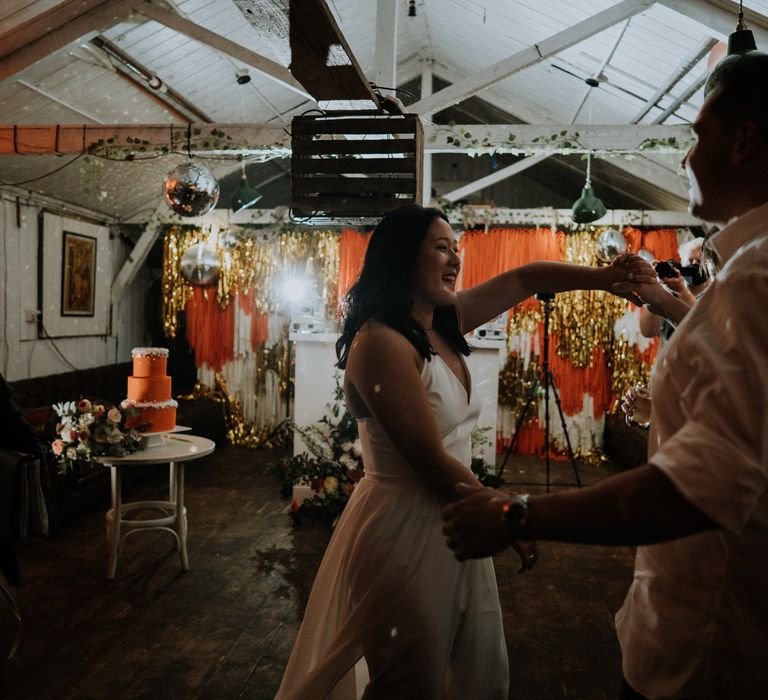 Bride and groom dancing with a orange, white and gold tassel wall in the background 