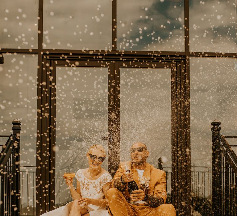 Bride and groom champagne popping moment with groom in a mustard suit and bride in a princess wedding dress smiling 