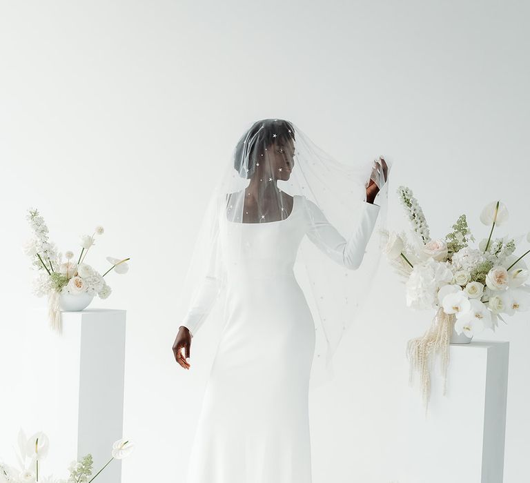 Black bride in a minimalist wedding dress with long sleeve wearing a star covered wedding veil standing neck to all white wedding flowers on plinths