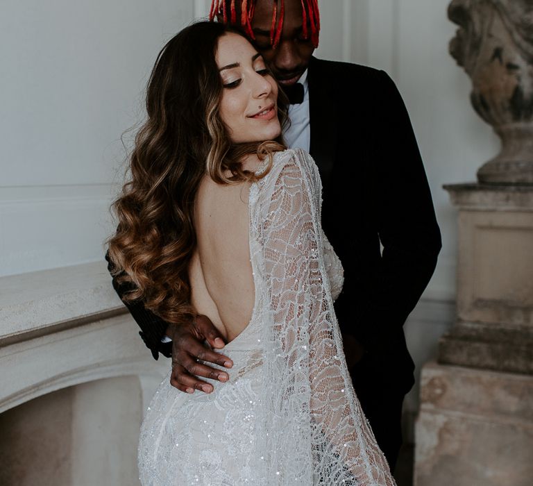 Bride in a sparkly lace wedding dress with low back and long wavy hair being embraced by her groom in a black velvet jacket at Bylaugh Hall 