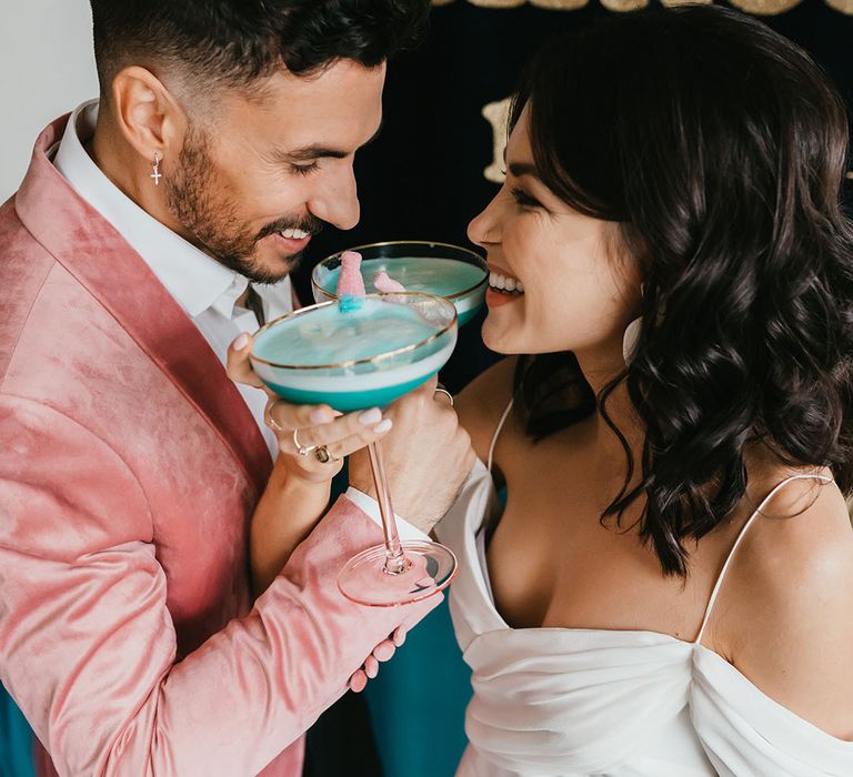 Groom in a pink jacket and bride in a cold shoulder wedding dress drinking blue cocktails with sweets in 