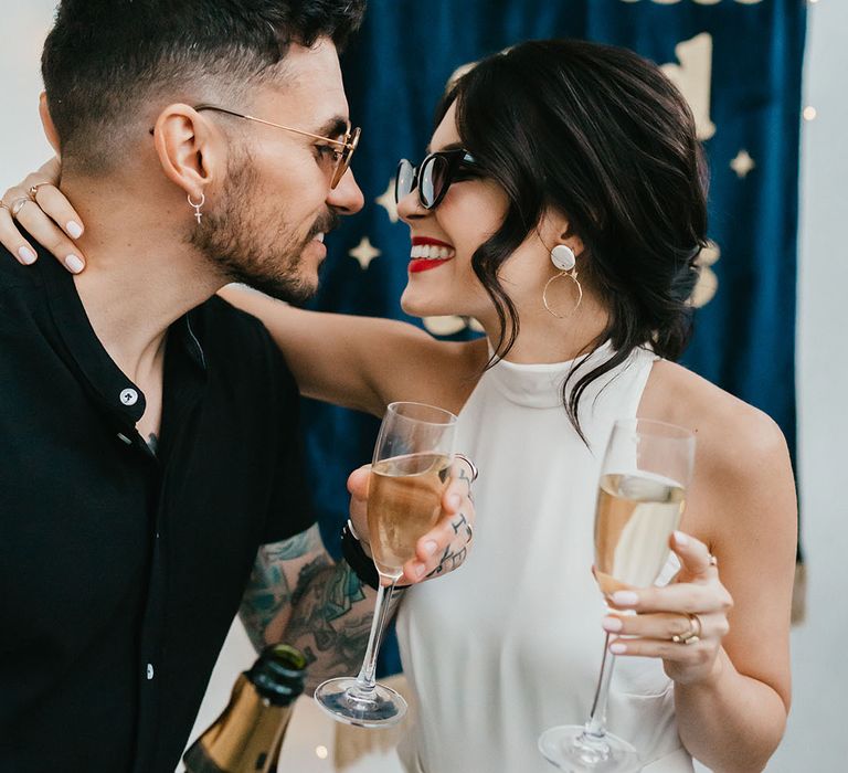 Bride with pinned up hair wearing sunglasses, hoop earrings and red lipstick smiling at her groom in a black shirt whilst they sip champagne 