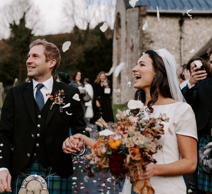 Bride in veil and groom in kilt delight in falling confetti