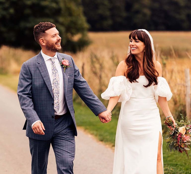 Groom in a blue check suit holding hands with his bride in a strapless wedding dress with lace puff sleeves and front split design 