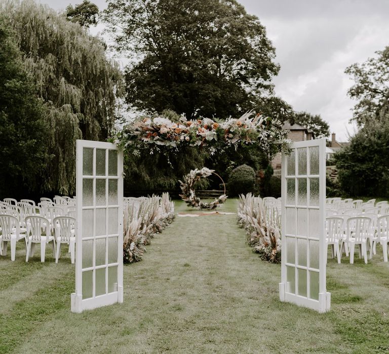 Green glass doors and floral garland open onto garden wedding ceremony with white chairs, pampas grass floral installations and floral Moorgate for DIY garden wedding