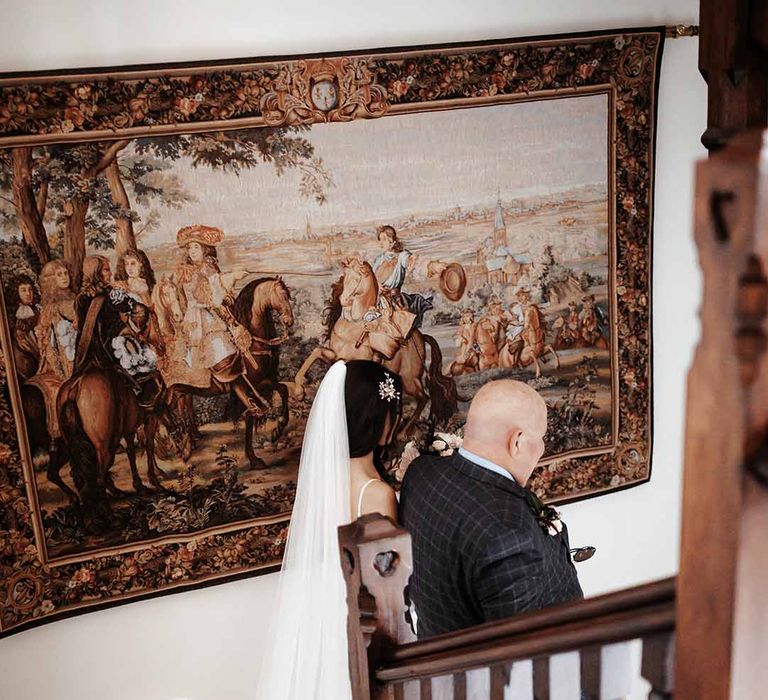 Brides long wedding dress flows as she walks down the stairs in country manor with her dad 