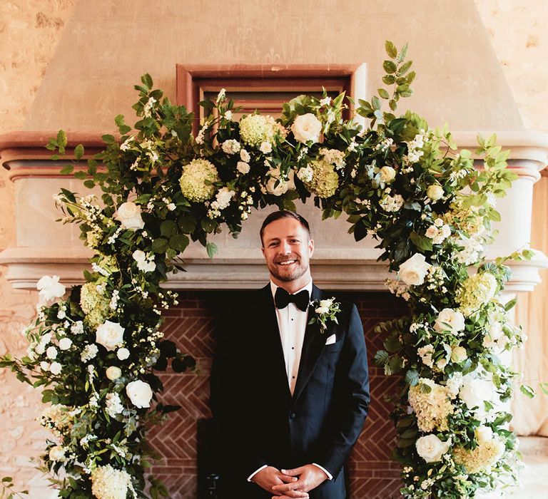 Groom stands in front of floral archway 