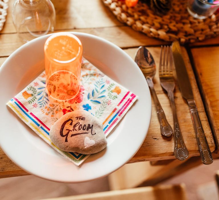 White plate with colourful napkin, orange cup and pebble table place names for rustic wedding reception