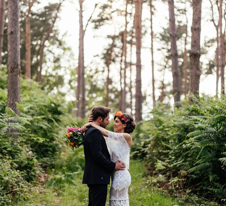 Bride in off the shoulder Grace Loves Lace wedding dress and flower crown holding colourful wedding bouquet hung groom in ball suit in woodland after outdoor wedding ceremony