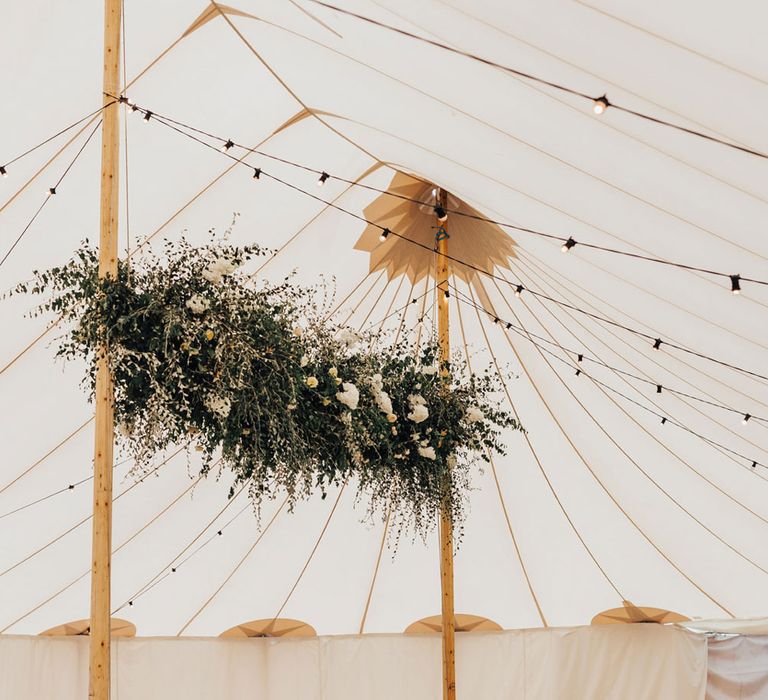 Long wooden tables with amber glassware and florals with large flower cloud and hanging bulb lights for tent wedding reception in Hertfordshire