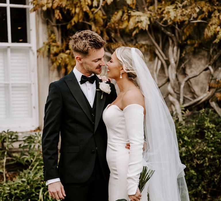 Groom in a black three-piece suit and shiny shoes embracing his bride in a fitted mermaid train wedding dress and cathedral length veil in front of Northbrook Park wedding venue 