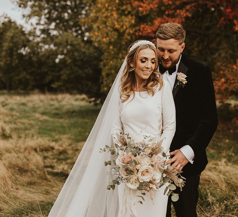 Groom in a tuxedo embracing with his bride in a long sleeve wedding dress, pearl headband as she holds a pink rose bouquet 