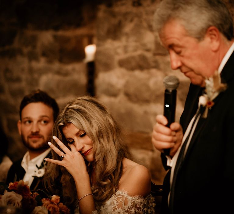 Emotional bride in boho wedding dress wipes her eyes whilst man in dark suit and floral buttonhole holds microphone whilst making a speech 