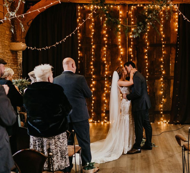 Bride in tulle wedding dress with train and veil kisses groom in dark suit in front of fairy lights for barn wedding ceremony