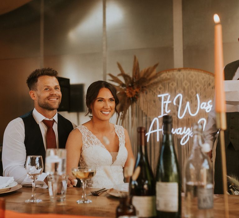 Smiling bride in lace wedding dress sits with groom in burgundy tie and waistcoat as guest makes speech during barn wedding reception with rustic orange decor