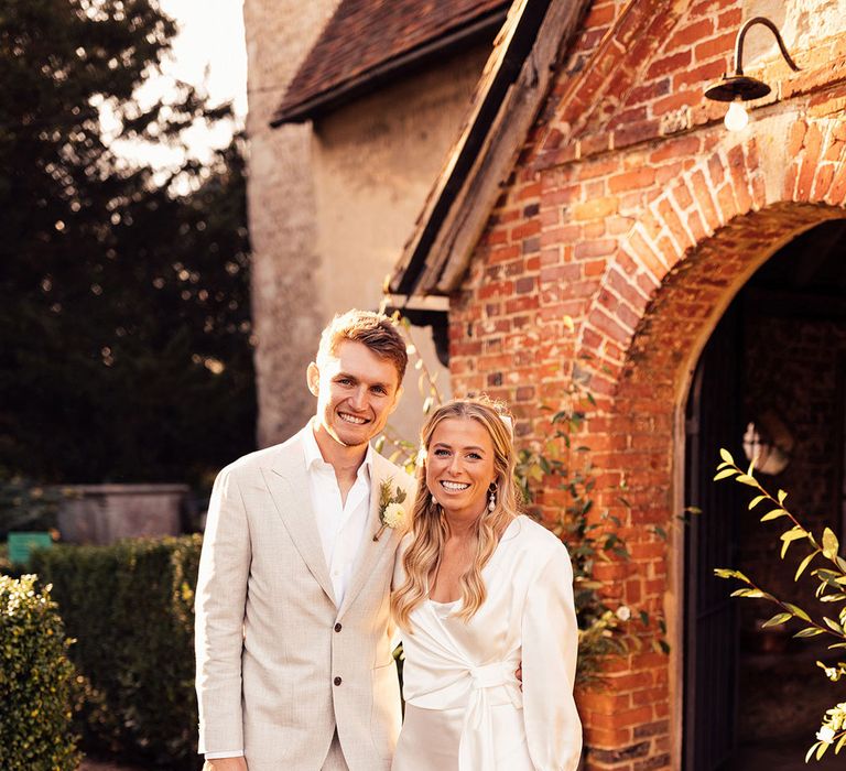 Groom in light grey suit with open white shirt and yellow floral buttonhole stands outside church with bride in satin bridal outfit holding pink and yellow mixed wedding bouquet 