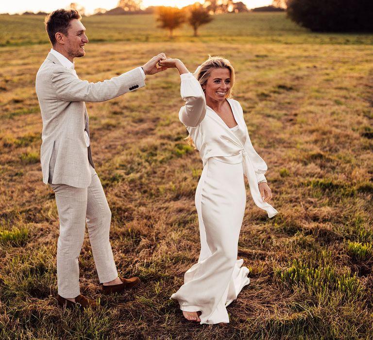 Bride in white The Own Studio satin dress with wrap top dances in field with groom in light grey suit 