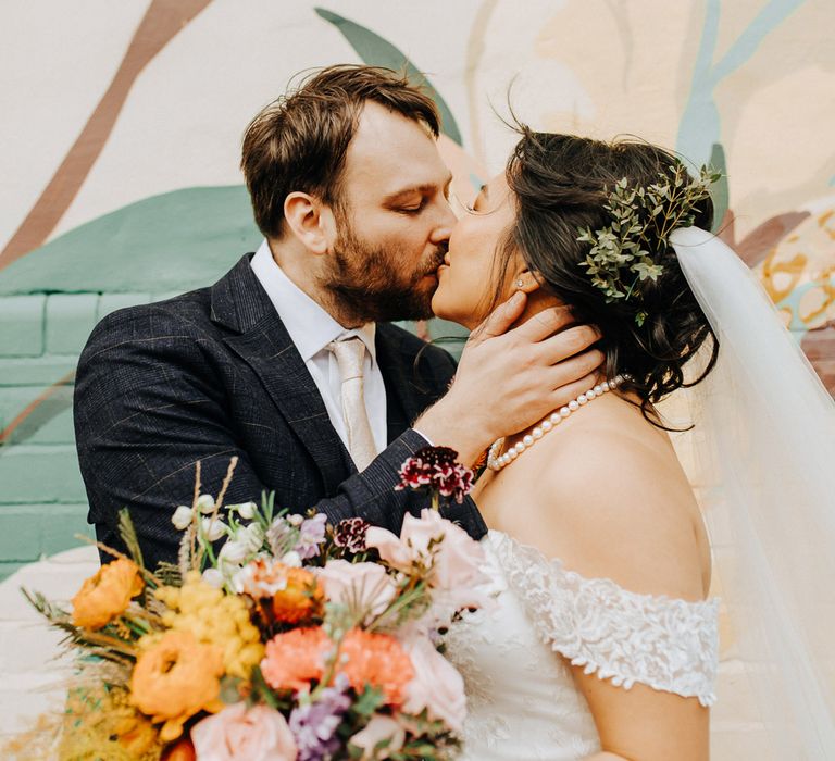 Groom in blue check suit with light pink tie kisses bride in off the shoulder lace dress and pearl necklace holding orange, yellow and pink bouquet by street art at Scale in Liverpool