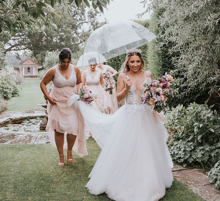 Bridesmaids walk with the bride in the rain with clear umbrellas to the wedding ceremony 