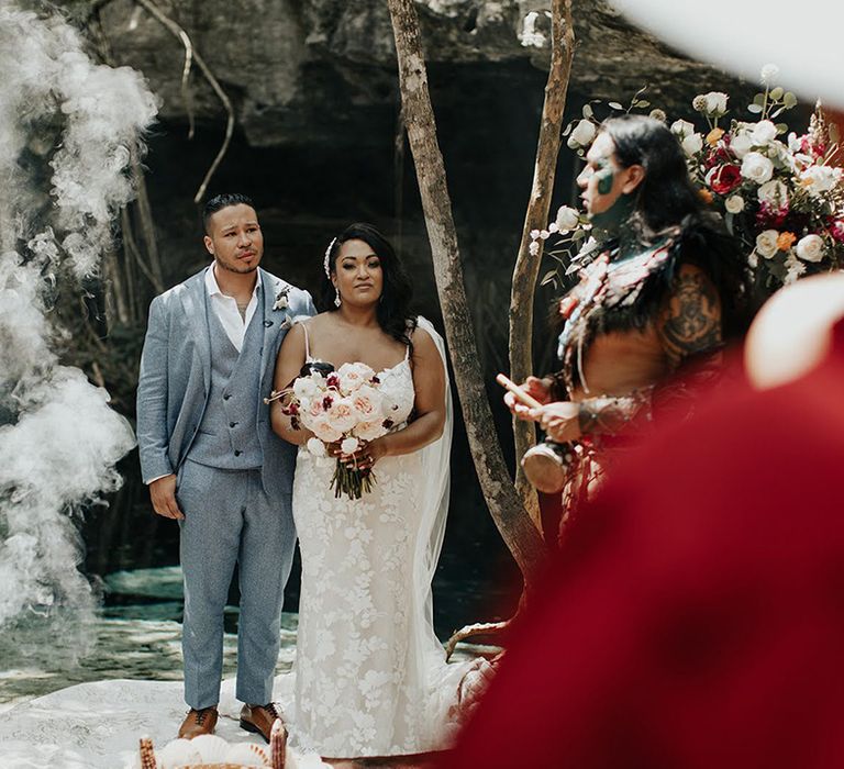 Bride in lace floral wedding gown and bridal hair piece accessory stands with groom at Mayan wedding ceremony