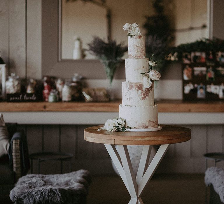 Four-tier wedding cake with white, pink, gold frosting and white rose flower decorations at barn venue