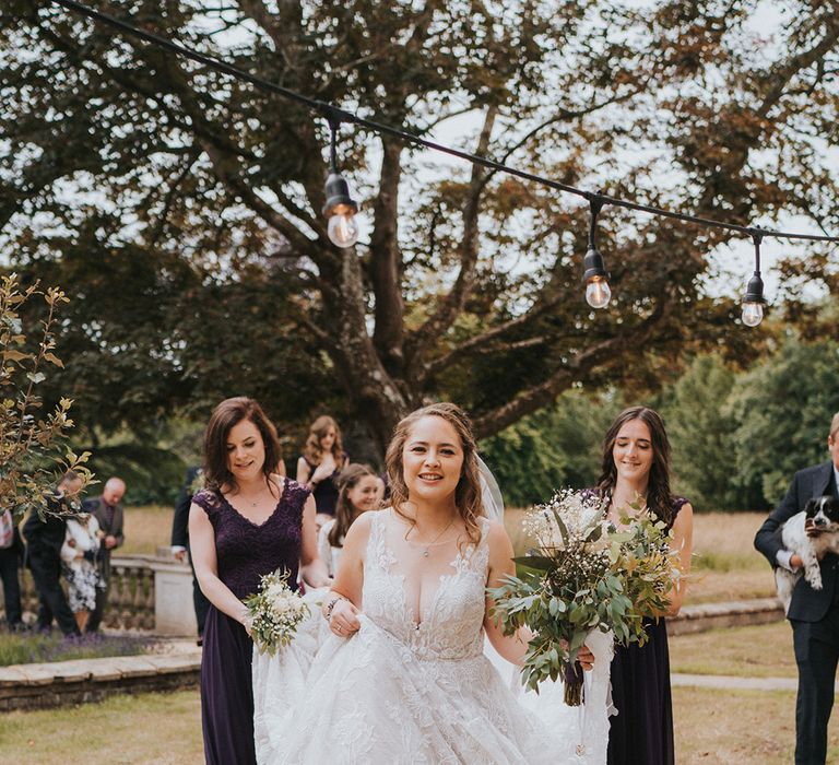 Bridesmaids in deep purple lift up the birde's lace dress as she walks with pearl Charlotte Mills wedding shoes