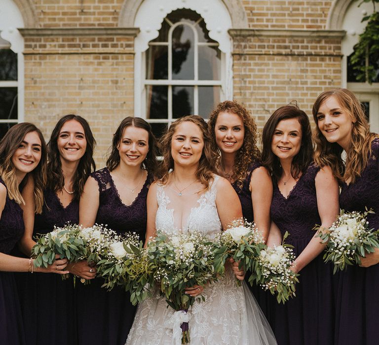 Bride in lace illusion neck wedding dress with bridesmaids in deep purple lace dresses holding gypsophila and rose bouquets with lavender 