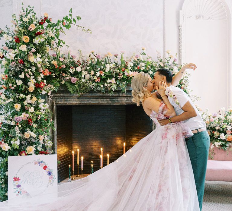 LGBTQI+ couple in a pink flower wedding dress and white t-shirt kissing in front of a fireplace decorated with colourful flowers and candles 