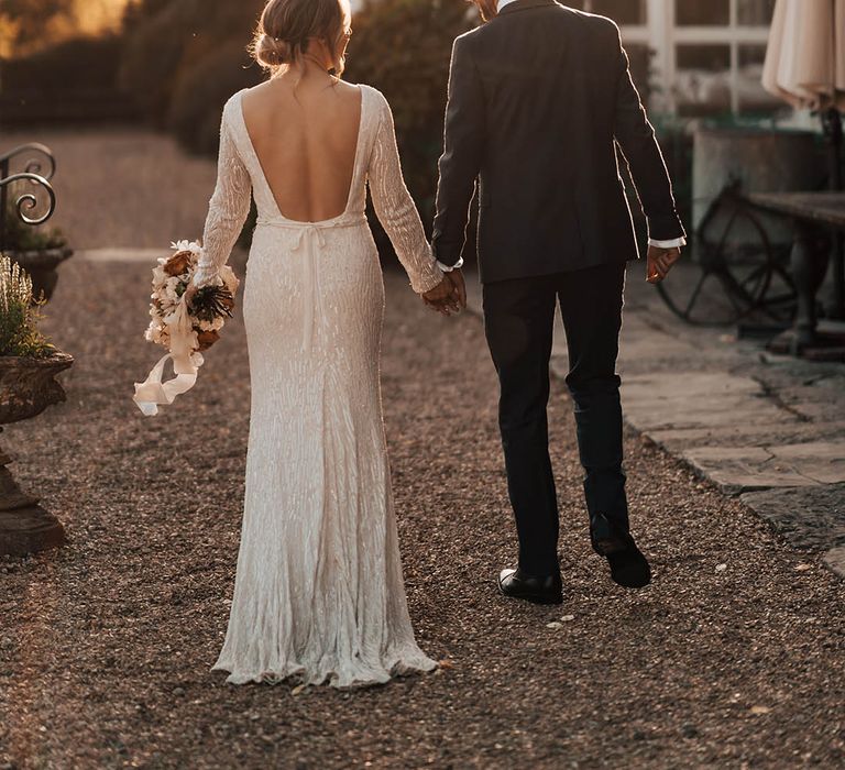 Bride in beaded vintage style wedding dress with low back holds hands with groom as they walk outside their wedding venue