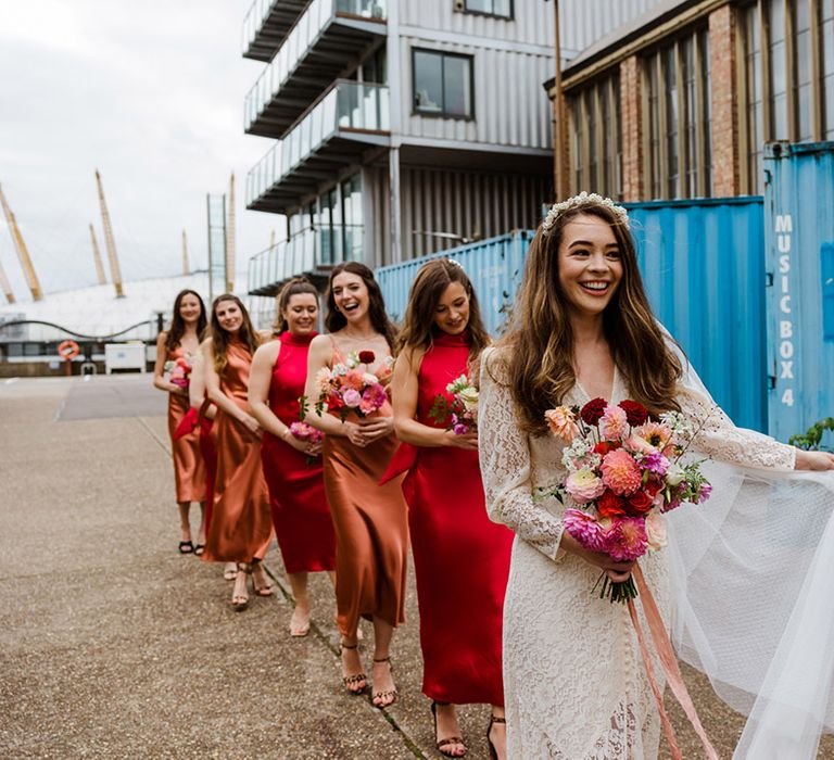 Bride in lace wedding dress with buttons and pearl headband leads bridesmaids in orange and red satin dresses to the wedding in London